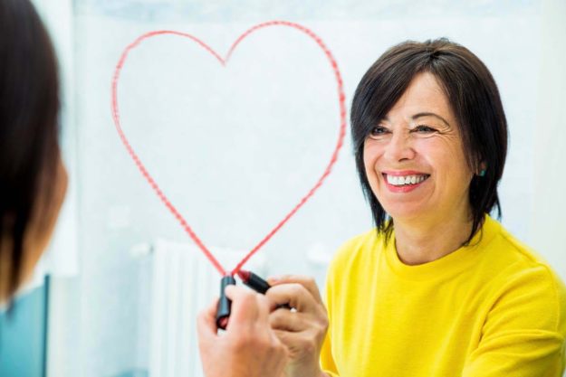 a woman draws a lipstick heart around her reflection in the mirror