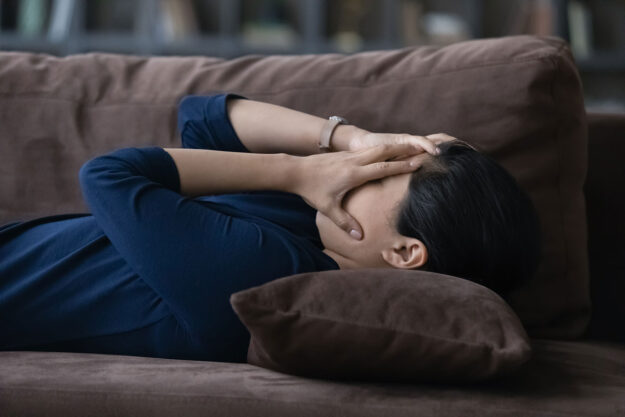 a person lays on a couch and puts their hands over their face during the physical symptoms of anxiety