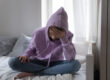 a kid sits on a bed in a hoodie showing signs of cutting