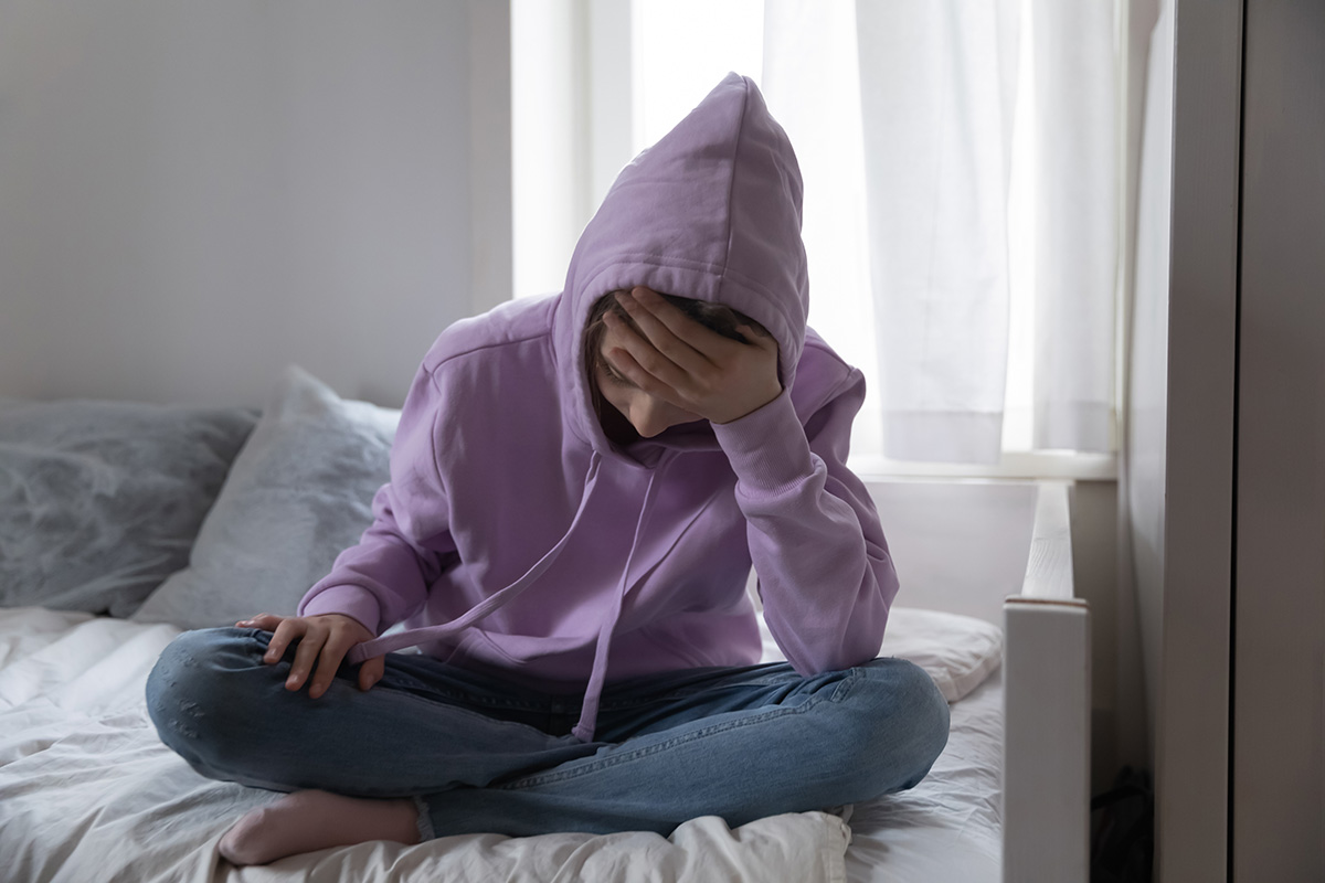 a kid sits on a bed in a hoodie showing signs of cutting