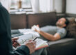 a person lays on a couch in the background while in the foreground a therapist takes notes during mental health care treatment