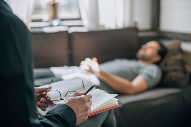 a person lays on a couch in the background while in the foreground a therapist takes notes during mental health care treatment