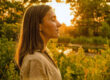 A woman with her eyes closed stands in profile, lit by warm golden sunlight among green foliage.