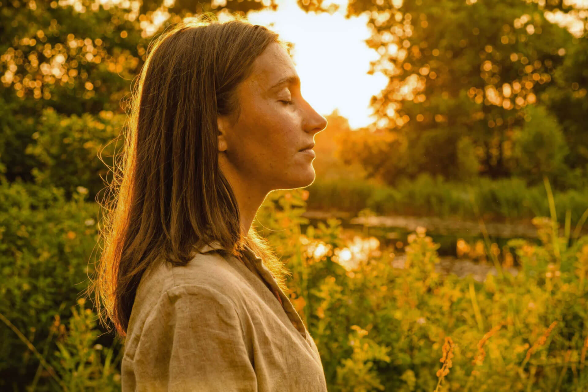 A woman with her eyes closed stands in profile, lit by warm golden sunlight among green foliage.