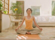 A woman sits cross-legged on a yoga mat with her eyes closed in a bright, plant-filled room.