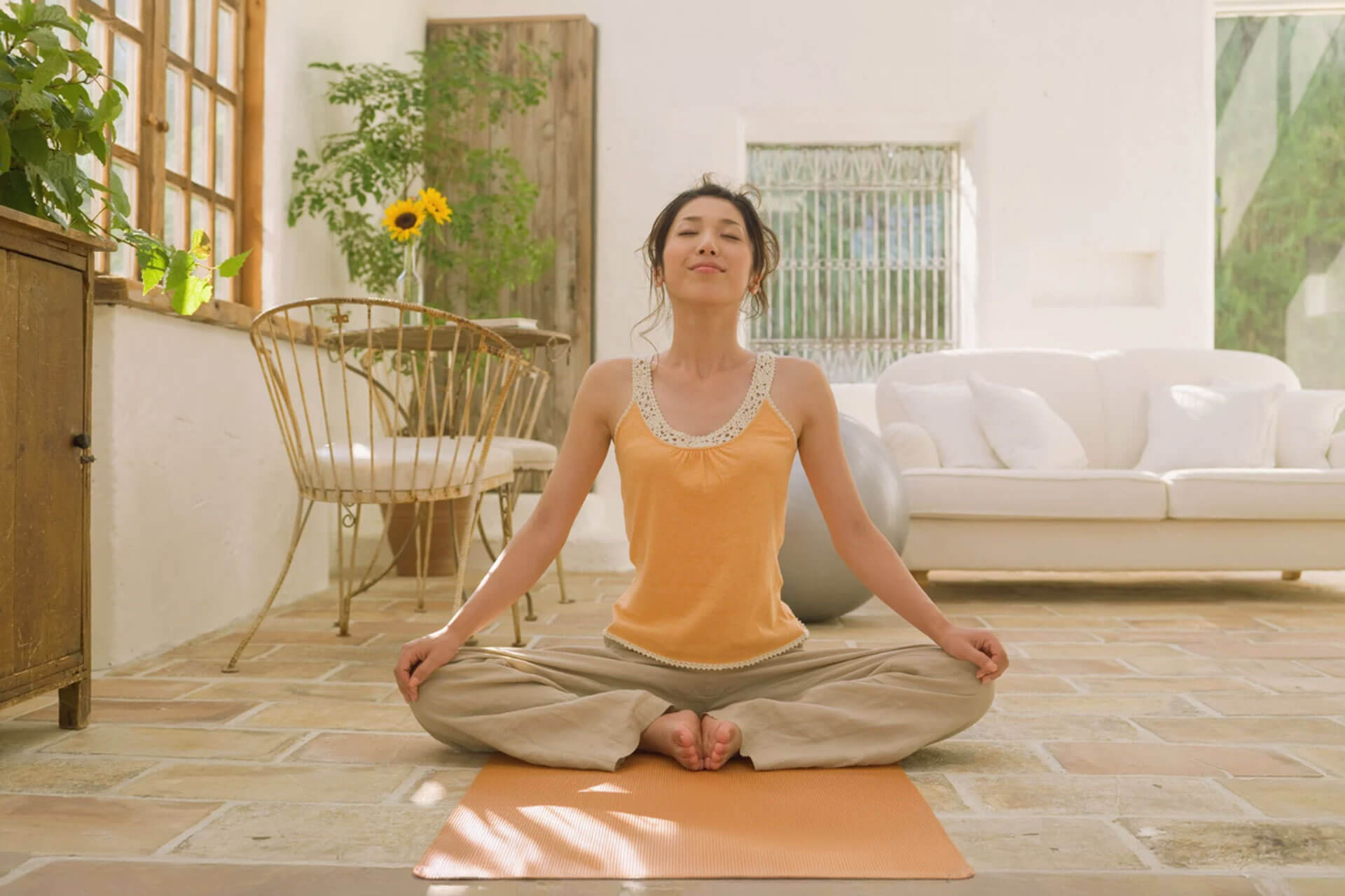 A woman sits cross-legged on a yoga mat with her eyes closed in a bright, plant-filled room.