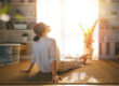 A woman sits on a woven rug with her face tilted toward a sunlit window in a cozy living room.
