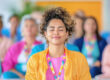 A smiling woman in a yellow jacket and colorful lanyard closes her eyes with a blurred group behind her.