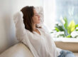 A relaxed woman leans back on a couch with her hands behind her head and her eyes closed.