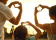 A man, woman, and child make heart shapes with their hands against a glowing sunset sky.