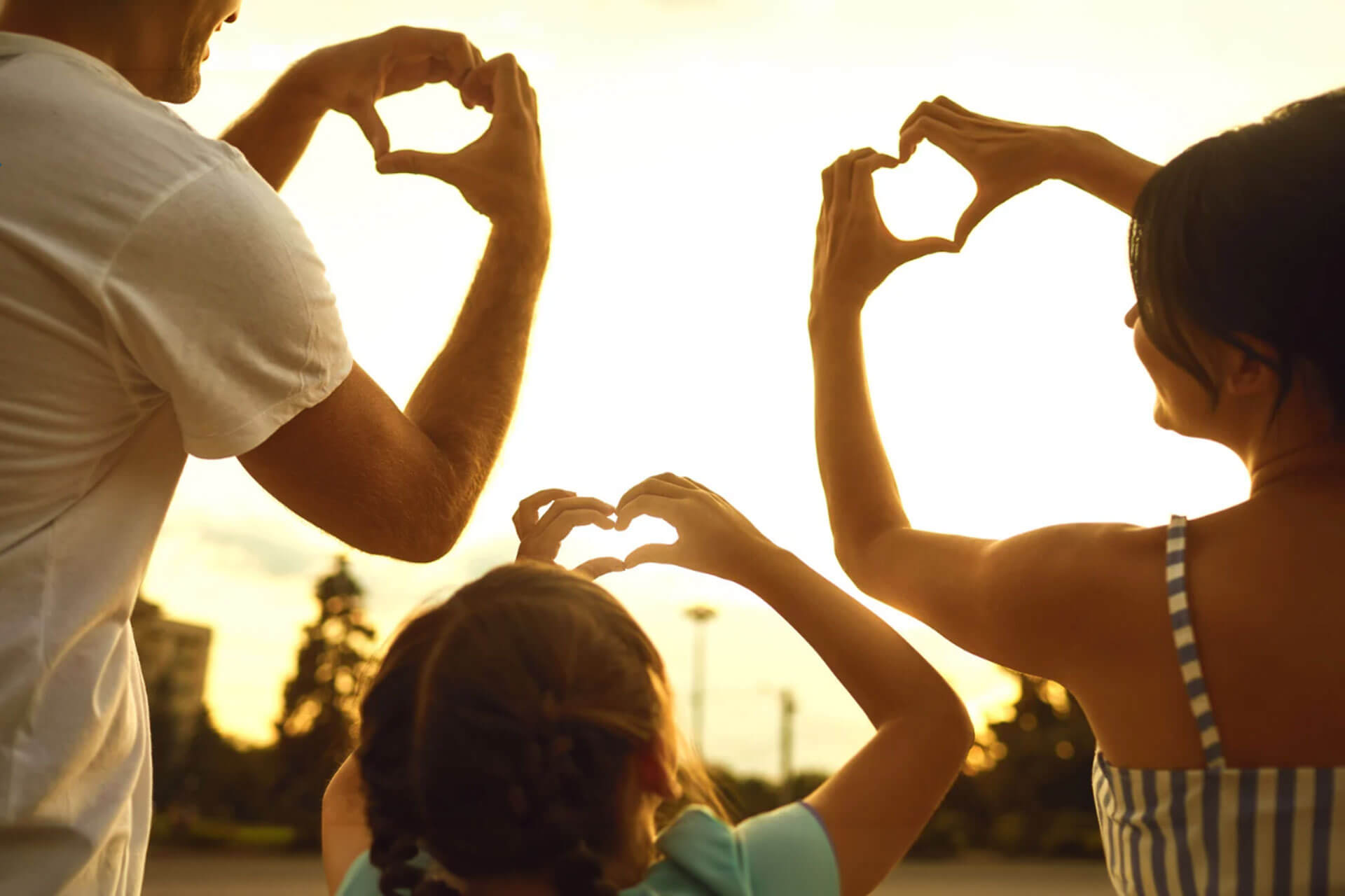A man, woman, and child make heart shapes with their hands against a glowing sunset sky.