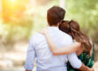 A woman rests her head on a man's shoulder as they embrace and face a sunlit, blurred background.
